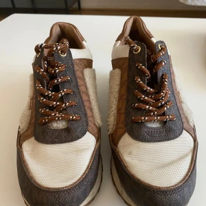 Michael Kors sneakers med gulddetalj - Snygga sneakers från Michael Kors i beige och brunt med gulddetalj på hälen. Skorna har mönstrad logotyp, snörning och inslag av mjuk teddy på sidorna. Materialmix av skinn, syntet och teddy ger en unik look. Perfekta för dig som gillar statement-skor med lyxig vibe.