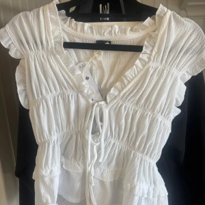Vit blus med volanger från Gina Tricot - Supersöt vit blus från Gina Tricot i storlek XS. Blusen har volanger vid axlar och nederkant, v-ringning med snörning framtill och ett lätt krinklat tyg. Perfekt för dig som gillar romantisk och trendig stil med extra detaljer.