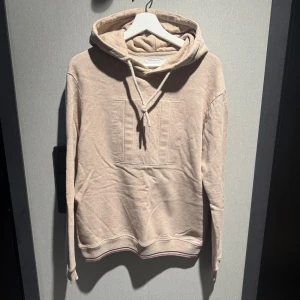 Beige hoodie från Les Deux - Snygg beige hoodie från Les Deux med broderad logga på bröstet och klassisk huva med snörning. Tröjan har långa ärmar och ribbad mudd med diskret röd och blå rand längst ner. Mjuk insida i bomullsmix, tröjan är sparsamt använd och i nyskick. 