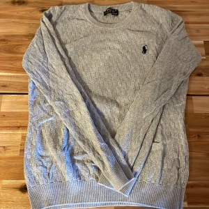 Grå stickad tröja från Polo Ralph Lauren - Snygg grå stickad tröja från Polo Ralph Lauren med diskret strukturmönster och klassisk svart logga broderad på bröstet. Rund hals och långa ärmar med ribbade muddar för en clean look. Perfekt för dig som gillar stilrena och tidlösa plagg.