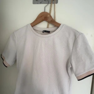 Vit ribbad t-shirt från Zara - Snygg vit ribbad t-shirt från Zara med rund hals och korta ärmar. Ärmsluten har en beige kant med svart detalj som ger en cool kontrast. Perfekt för dig som gillar stilrena och enkla plagg med en twist.