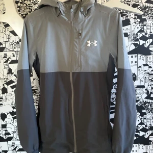 Grå vindjacka från Under Armour - Hejsan, säljer just nu en snygg tvåfärgad vindjacka från Under Armour i ljusgrått och mörkgrått. Jackan är i storlek xs och har huva, dragkedja hela vägen och en bröstficka med dragkedja. 