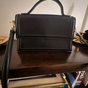 Svart handväska från Gina Tricot - Stilren svart handväska från Gina Tricot i skinnimitation med raka linjer och guldfärgade detaljer. Väskan har både handtag och axelrem, samt ett fack med dragkedja på insidan. Använd ett fåtal gånger men i fint skick. 