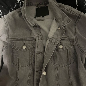Grå jeansjacka med knappar - Snygg grå jeansjacka i klassisk modell med silverfärgade knappar framtill och på bröstfickorna. Jackan har lång ärm, två bröstfickor med lock och markerade sömmar som ger en cool vibe. Perfekt att slänga över en hoodie eller t-shirt 