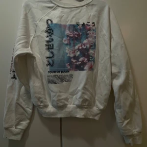 Vit sweatshirt med Japan-print - Den är i mycket bra skick men skrynklig. Vit sweatshirt med coolt tryck på ryggen och ärmen, inspirerat av Japan och körsbärsblommor. Trycket har japanska tecken och texten 'TOUR OF JAPAN'. Tröjan har rund hals och långa ärmar, perfekt för dig som gillar streetstyle och unika prints.