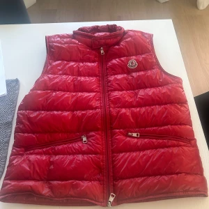 Röd dunväst från Moncler - Snygg röd dunväst från Moncler med quiltad design och hög krage. Västen har två praktiska fickor med dragkedja och ett broderat Moncler-märke på bröstet. Materialet är glansigt och känns lyxigt, perfekt för att lyfta din outfit under kyliga dagar.                                                                             Storlek: 3