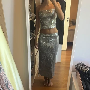 Silvrig glittrigt sett - Säljer ett silvrigt sett med glittriga paljetter från Topshop. Toppen är axelbandslös och har en omlott-design upptill som ger en snygg och modern look. Materialet är syntet med paljettöverdrag, storlek 36-38 på både topp och kjol. Perfekt för temafester eller liknande!!                    Inköpspris: 600 