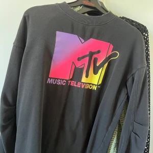Svart MTV sweatshirt med färgglad logga - Cool svart sweatshirt med stor MTV Music Television-logga i lila, rosa och gul på bröstet. Tröjan har rund hals, långa ärmar och är gjord i mjuk bomull. Perfekt för dig som gillar retro och streetstyle.