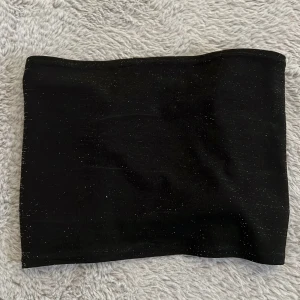 Svart glittrig bandeau tubtopp med snörning - Säljer en svart bandeau tubtopp med glittriga detaljer och snygg snörning baktill. Toppen är axelbandslös och har en cool, croppad passform som verkligen sticker ut. Perfekt för dig som vill ha något extra till outfiten.
