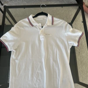 Vit polo t-shirt från Prada - Vit polo t-shirt från Prada med klassisk krage och knappar framtill. Röd och svart randig detalj på krage och ärmslut ger en clean vibe. Diskret Prada-logga på bröstet. Tillverkad i mjuk bomull som känns skön mot huden. Perfekt för dig som gillar stilren design. Kommer inte till användning mer perfekt skick 