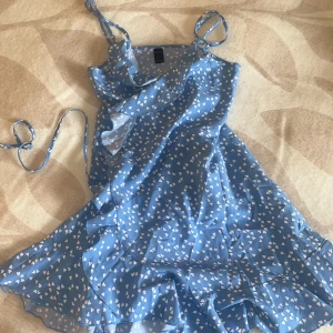 Blå hjärtmönstrad kortklänning - Supersöt blå kortklänning från SHEIN med vita hjärtan över hela tyget. Klänningen har tunna axelband, volangdetaljer och omlottkänsla framtill. Perfekt för sommarens alla tillfällen, lätt och luftig i syntetmaterial.