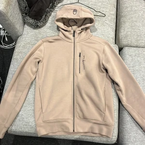 Beige hoodie från Sail Racing - Snygg beige hoodie från Sail Racing med hel dragkedja och huva. Tröjan har en bröstficka med dragkedja och broderad logga på framsidan. Insidan är mjuk och fleecefodrad, perfekt för kyliga dagar. Långärmad och med en clean, stilren look. Har en fläck på högra underarmen. Kan säkert tvättas bort.