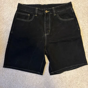 Svarta Carhartt jeansshorts, storlek 32 - Svarta jeansshorts från Carhartt med kontrasterande vita sömmar och klassisk femficksdesign. Shortsen har normal passform(S/M), knapp och dragkedja framtill samt Carhartt-logga på bakfickan. Perfekta för dig som gillar streetwear och robust stil.