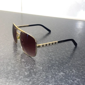 Louis Vuitton guldiga solglasögon - Snygga solglasögon från Louis Vuitton med fyrkantiga linser i mörkröd ton. Bågarna är guldiga med svarta skalmar och har ett randigt mönster på sidorna. Metallramen ger en lyxig känsla och designen är riktigt stilren.