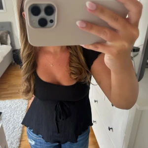 Svart blus med knyt och volang - Supersöt svart blus med korta ärmar, knyt i midjan och volangkant nertill. Toppen har en fyrkantig ringning och är tillverkad i ett lätt, lite strukturerat tyg som ger en skön och luftig känsla. Perfekt att styla med jeans eller kjol för en trendig look.