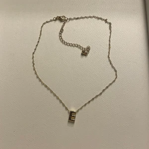 Guldigt halsband med E-berlock & ringar - Säljer ett tunt guldigt halsband med en stilren E-berlock samt tre matchande ringar i guldton. En ring har ett snirkligt mönster, en är slät och en har en liten rund platta. Perfekt om du gillar enkla och stilrena smycken. Ringarna är köpta i stl XS/S från H&M eller Gina
