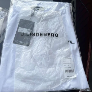 Vit t-shirt från J.Lindeberg XL - Fräsch vit t-shirt från J.Lindeberg i storlek XL med svart logotyp tryckt över bröstet. Klassisk passform och rund hals. Perfekt för dig som gillar stilrena och enkla plagg med tydlig branding.