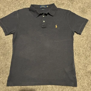 Mörkblå Ralph Lauren piké  storlek M - Riktig bra skick Snygg mörkblå t-shirt från Polo Ralph Lauren i slim fit-modell. Klassisk pikékrage med två knappar och korta ärmar. Ikonisk gul broderad logga på bröstet. Tillverkad i mjuk bomull som känns skön mot huden.