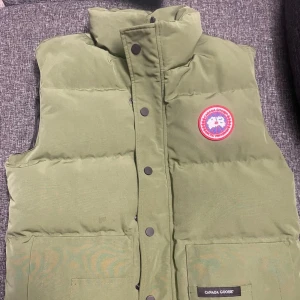 Grön Dunväst  - Säljer en grön dunväst från Canada Goose med hög krage och två stora fickor framtill. Västen har klassisk logga på bröstet och stängs med tryckknappar samt zip. Perfekt för lager-på-lager och riktigt snygg till streetwear-stilen.