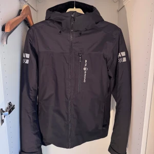 Svart vindjacka från Sail Racing - Svart vindjacka från Sail Racing med huva och dragkedja framtill. Jackan har en bröstficka med vertikal dragkedja och logga på bröstet samt tryck på båda ärmarna. Perfekt för blåsiga dagar och snygg till streetwear.