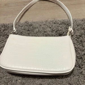 Vit axelväska med krokopräglat mönster - Snygg vit axelväska med krokopräglat mönster och stilren form. Väskan har en bred axelrem och stängs med dragkedja upptill. Perfekt storlek för det viktigaste och ger en clean vibe till din outfit.