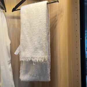Vit fluffig halsduk - Supermjuk och fluffig vit halsduk med stilrena fransar i kanten. Perfekt storlek för att svepa runt halsen och få en cozy look. Materialet är tjockt och värmande, perfekt för kyliga dagar. Enkel design som passar till de flesta jackor.