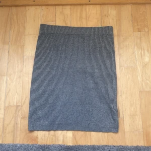 Grå ribbad kortkjol - Snygg grå ribbad kjol med rak passform och kort längd. Har dock ett pyttelitet hål men syns inte. Kjolen är stickad och har en enkel, stilren look som passar perfekt till en chill outfit. Materialet är mjukt och följsamt, vilket gör den skön att bära. Perfekt att matcha med en oversized hoodie eller t-shirt.