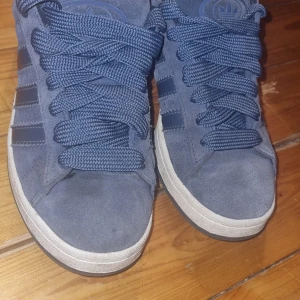 Blå adidas sneakers i mocka - Säljer ett par snygga blå adidas sneakers med klassiska tre ränder på sidan och logga på tungan och hälen. Skorna är gjorda i mjuk mocka med matchande blå snören och vit sula. Perfekt för dig som gillar stilrena och sportiga vibes.