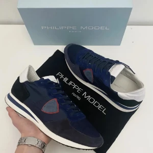 Blå Philippe Model sneakers - Skick 9/10 endast testade någon gång . Storlek 45 .påse , box och prislapp följer med vid köp. Snygga sneakers från Philippe Model i marinblått med vita och svarta detaljer. Skorna har snörning, rund tå och en platt sula. Materialmix av mocka och syntet, med en cool röd kant runt loggan på sidan. Perfekta för dig som gillar stilrena och sportiga skor.pytte lite skumgummi som lossnat vid hälarna men inget som påverkar 