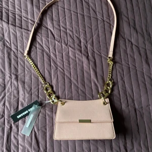 Ljusrosa axelväska från Like Dreams - Snygg ljusrosa axelväska från Like Dreams med croco-mönstrad yta och guldfärgade kedjedetaljer. Väskan har ett stilrent lock med magnetknäppning och en justerbar axelrem. Perfekt storlek för det viktigaste och ger en trendig touch till din outfit.