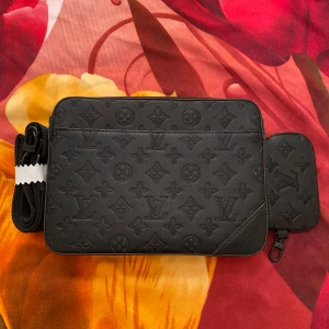 Svart Axelväska  - Säljer en svart axelväska med klassiskt monogrammönster i skinn. Väskan har en rektangulär form, justerbar axelrem och en matchande liten plånbok som kan fästas på väskan. Perfekt för dig som gillar stilren och lyxig design.