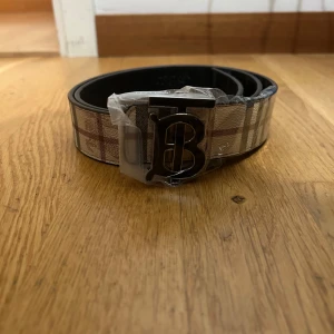 Bälte med två färger med burberry tag - Snyggt bälte från Burberry med klassiskt rutigt mönster i beige, brunt och rött. Bältet har ett stort metallspänne med ett stilrent B i silverfärg. Perfekt accessoar för att lyfta din outfit med en lyxig touch.