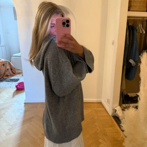 Grå stickad hoodie med huva - Mysig grå stickad hoodie med breda ribbade muddar och huva. Tröjan har snörning vid halsen och en loose passform som ger en chill vibe. Perfekt för höst och vinter när man vill vara både bekväm och snygg.