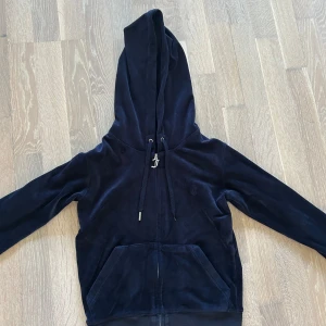Mörkblå velour hoodie från Juicy Couture - Säljer en mörkblå hoodie från Juicy Couture i mjuk velour med dragkedja och huva. Tröjan har långa ärmar, snörning i huvan med metalländar och ett snyggt J-emblem både på dragkedjan och broderat på bröstet. Perfekt för chill dagar.