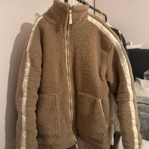 Beige teddyjacka med vita detaljer - Mysig teddyjacka i beige med vita ränder längs ärmarna och dragkedja framtill. Jackan har två stora fickor och hög krage som håller dig varm. Perfekt för kyliga dagar och har en avslappnad, loose passform.