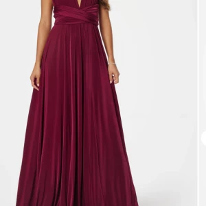 Vinröd långklänning med omlott - Säljer en vinröd långklänning med omlott och v-ringad hals. Klänningen har ett snyggt fall och breda axelband, samt ett knyt i midjan som ger en elegant siluett. Materialet är mjukt och har en lätt glans, perfekt för bal eller fest.