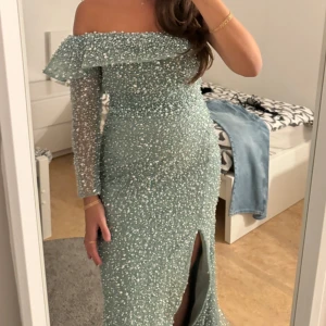 Glittrig ljusblå långklänning med slits - Säljer en ljusblå långklänning med glittriga pärlor över hela tyget. Klänningen har offshoulder-design, transparenta långärmar och en snygg slits framtill. Passformen är slim och tyget känns lyxigt och festligt. Perfekt för bal eller bröllop!