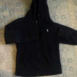 Svart hoodie från Polo Ralph Lauren som är använd ett par gånger bara  - Svart hoodie från Polo Ralph Lauren med dragkedja och huva. Klassisk vit Polo-logga på bröstet, snörning i huvan och två fickor framtill. Perfekt för chill dagar och enkel att matcha med jeans eller joggers.