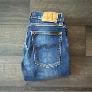 Blå skinny jeans från Nudie Jeans - Säljer ett par klassiska blå skinny jeans från Nudie Jeans med snygga slitningar och kontrastsömmar. Jeansen har fem fickor, normal midja och är tillverkade i stretchig bomullsdenim för en bekväm passform. Perfekta för dig som gillar en smal siluett.