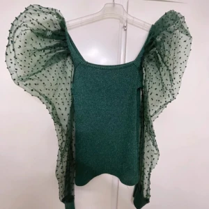 Grön glittrig topp med puffärmar - Unik grön topp med glittrigt ribbat tyg och stora puffärmar i transparent mesh med prickigt mönster. Ärmarna ger en dramatisk look och toppen har en croppad passform. Perfekt för dig som vill sticka ut och älskar coola detaljer.