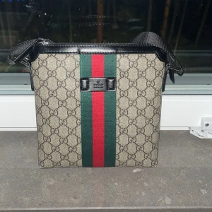 Gucci axelväska med GG-mönster - Snygg axelväska från Gucci med klassiskt GG-mönster i beige och brunt. Väskan har en bred grön och röd rand i mitten, svarta skinn-detaljer och justerbar axelrem. Insidan är fodrad i brunt tyg och har en dragkedja upptill. Perfekt för dig som vill ha en ikonisk designer-väska.