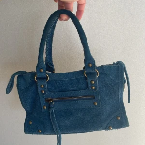 Blå mocka handväska från lyckliga jag - Snygg blå handväska i mocka med coola metalldetaljer och dragkedja framtill. Väskan har två handtag med flätad look och två fack inuti där man får plats med mycket! Jag köpte väskan för 1500kr på lyckliga jag, men har bara använt den 2-3 gånger.