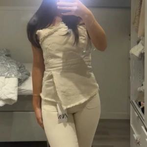 Beige tubtopp i jeans från Zara - Säljer en beige tubtopp från Zara i jeansmaterial med råa, fransiga kanter upptill och nedtill. Toppen har en asymmetrisk skärning framtill och coola sömdetaljer som ger en edgy vibe. Perfekt att styla med höga jeans eller kjol för en trendig look.