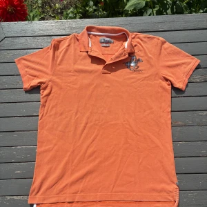 Orange pikétröja från Beverly Hills Polo Club - Snygg orange pikétröja från Beverly Hills Polo Club i slim fit-modell. Tröjan har klassisk krage, korta ärmar och ett stort rutigt polospelar-tryck på bröstet. Materialet är bomull och den har vita detaljer på insidan av kragen. Perfekt för en sportig och stilren look.