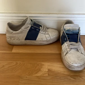 Valentino Open sneakers vit/blå - Säljer ett par Valentino Open sneakers i vitt skinn med bred blå rem över mitten och coola nitar längs hälen. Klassisk rund tå, platt sula och snörning framtill. Perfekt för dig som gillar lyxiga street vibes och vill sticka ut med designer på fötterna.