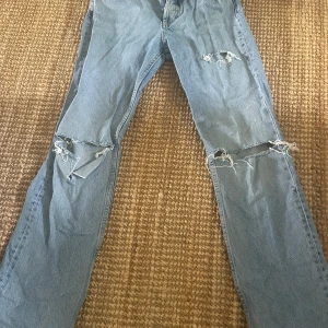 Blå midwaist straight jeans - Säljer ett par blå jeans med flera slitningar och hål på benen. Byxorna är från Zara och använda ett fåtal gånger, liknar de som säljs nu