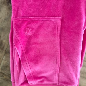 Juicy Couture mjukisset  - Rosa mjukisdress från Juicy Couture i mjuk velour med broderad JC-logga på fickan. Byxorna har bred resår i midjan och praktiska fickor, varav en med lock och silverfärgade knappar. Perfekta för chill eller streetwear.