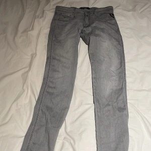 Replay Anbass grå slim jeans 28/32 - Snygga grå jeans från Replay, modell Anbass med slim fit. Klassisk femficksdesign och svarta detaljer på bakfickorna. Jeansen har raka ben och är tillverkade i mjukt jeansmaterial med en modern look. Perfekt för dig som gillar stilrena och coola jeans.