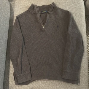 Grå stickad tröja från Polo Ralph Lauren - Snygg grå stickad tröja från Polo Ralph Lauren med half zip och långärmad design. Tröjan har en liten broderad logga på bröstet och ribbade muddar vid ärmslut och nederkant. Perfekt för dig som gillar klassisk stil med en modern touch. Passar mellan 150-160cm Inga fel jätte bra skick