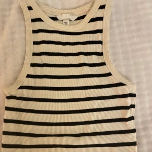 Randig croppad linnetopp från H&M - Säljer en fräsch croppad linnetopp från H&M i storlek XS. Toppen är vit med svarta horisontella ränder och har en ribbad struktur. Ärmlös design med rund halsringning, perfekt för dig som gillar en enkel men snygg look.